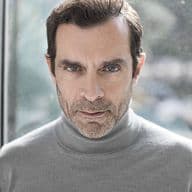 Φωτογραφία προσώπου ενός άνδρα με γαλανά μάτια και κοντά καστανά μαλλιά, που φοράει ένα γκρι ζιβάγκο πουλόβερ.