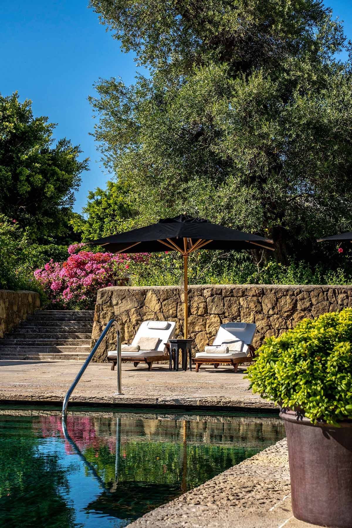 Two lounge chairs with cushions under a black umbrella by a pool, surrounded by lush greenery and vibrant flowers.