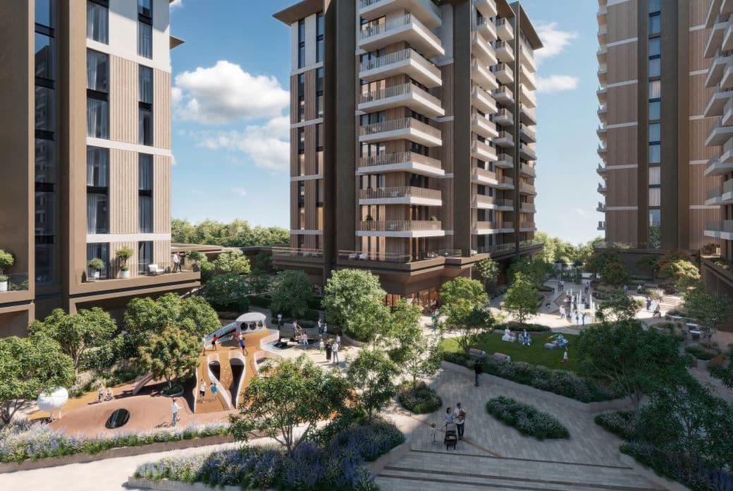 Modern residential complex with tall buildings, green landscaping, walking paths, and outdoor seating. Clear blue sky with scattered clouds above.