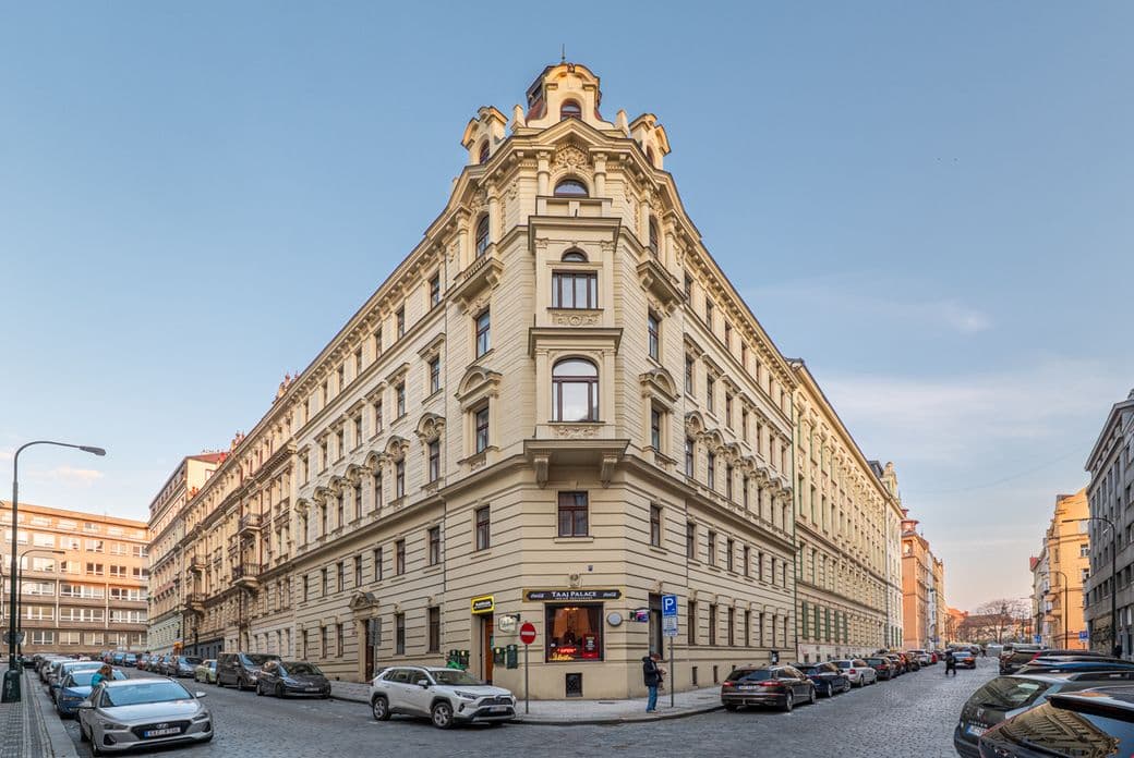 View of a multi-storey historic corner building with a light-coloured façade and rich decorative detailing. The structure is distinguished by an accentuated corner section topped with a tower-like roof feature. The façade includes stucco ornamentation, decorative cornices, and regularly arranged windows. The building stands at the intersection of two cobblestone streets lined with parked cars. Commercial premises occupy part of the ground floor, while the upper floors are residential. The image captures a classic urban setting with historic architecture under clear daylight conditions.