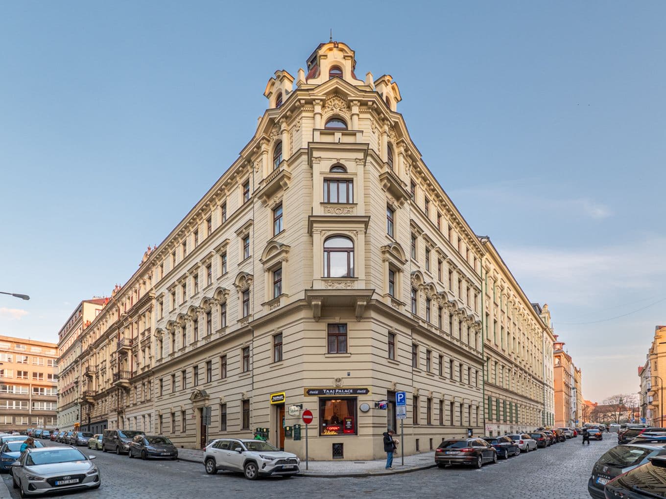 View of a multi-storey historic corner building with a light-coloured façade and rich decorative detailing. The structure is distinguished by an accentuated corner section topped with a tower-like roof feature. The façade includes stucco ornamentation, decorative cornices, and regularly arranged windows. The building stands at the intersection of two cobblestone streets lined with parked cars. Commercial premises occupy part of the ground floor, while the upper floors are residential. The image captures a classic urban setting with historic architecture under clear daylight conditions.