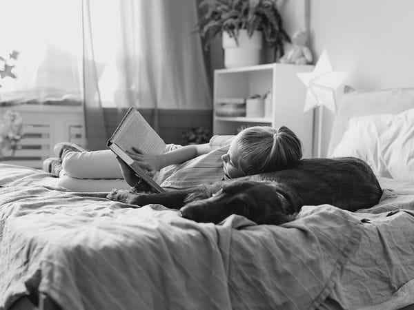 Ένα παιδί είναι ξαπλωμένο σε ένα κρεβάτι και διαβάζει ένα βιβλίο, ακουμπώντας το κεφάλι του σε ένα μεγάλο σκύλο. Το δωμάτιο έχει φυτά, ένα ράφι και φυσικό φως που μπαίνει από το παράθυρο. Η σκηνή φαίνεται ήρεμη και χαλαρή.