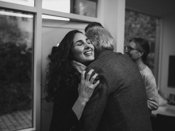 Μια γυναίκα χαμογελά με κλειστά μάτια ενώ αγκαλιάζει έναν ηλικιωμένο άνδρα μέσα σε ένα δωμάτιο. Μια άλλη γυναίκα στέκεται στο βάθος, μερικώς εκτός εστίασης. Η φωτογραφία είναι ασπρόμαυρη.