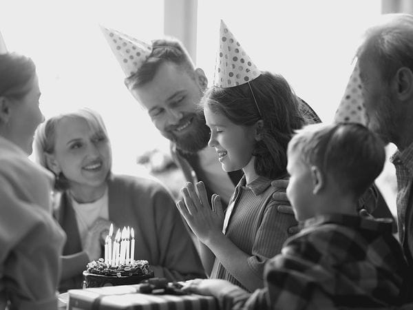 Μια ομάδα ανθρώπων με καπέλα για πάρτι περιβάλλει ένα χαμογελαστό κορίτσι, το οποίο κοιτάζει μια τούρτα γενεθλίων με αναμμένα κεριά. Η σκηνή παραπέμπει σε γιορτή γενεθλίων, με τα δώρα να είναι ορατά σε πρώτο πλάνο.