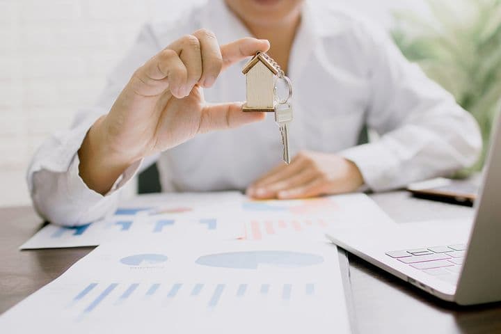 Person in a white shirt holding a small wooden house with keys attached, over a table with graphs and charts. A laptop is partially visible. The setting suggests a business or real estate context.