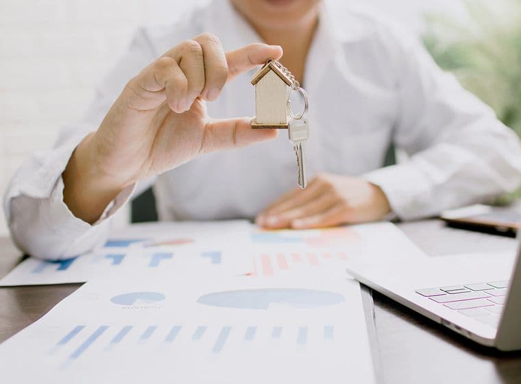 Person in a white shirt holding a small wooden house with keys attached, over a table with graphs and charts. A laptop is partially visible. The setting suggests a business or real estate context.