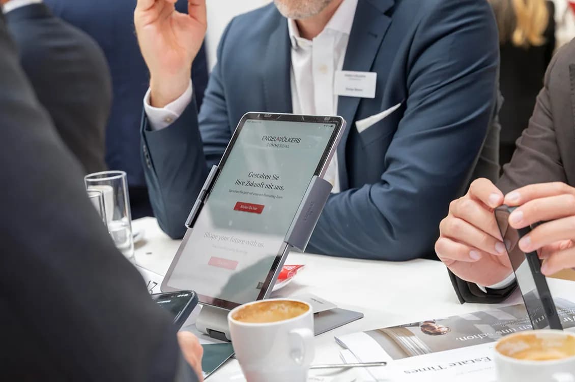 Geschäftsleute sitzen um einen Tisch herum; auf einem Ständer steht ein Tablet, auf dem „Engel & Völkers Commercial“ zu sehen ist, daneben stehen Kaffeetassen und Zeitschriften