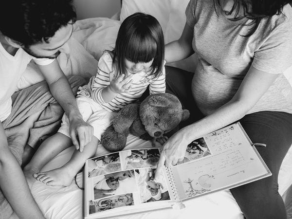 Ένα παιδί κάθεται ανάμεσα σε δύο ενήλικες σε ένα κρεβάτι και κοιτάζει ένα άλμπουμ με φωτογραφίες μωρών. Ένα λούτρινο αρκουδάκι βρίσκεται στην αγκαλιά του παιδιού και ο ένας ενήλικας φαίνεται να είναι έγκυος.