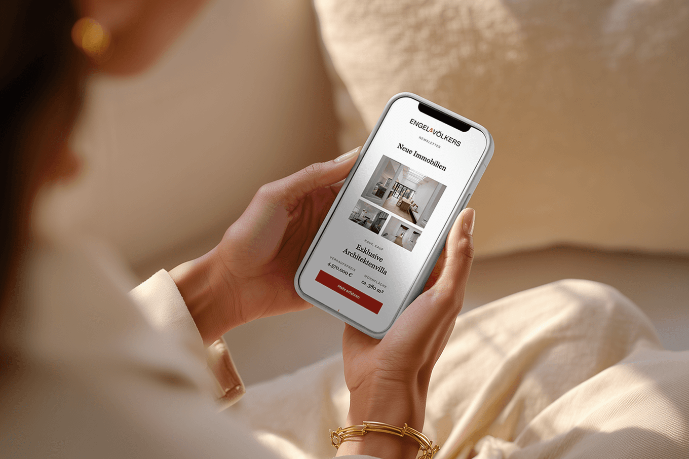 Person holding a smartphone displaying a real estate app with home listings, seated on a beige couch.