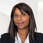 Aissatou Frisch-Baldé with long dark hair, wearing a black blazer and white blouse, stands smiling in front of a white background.