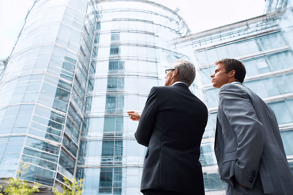 Two business professionals in suits stand outside, observing and pointing at a modern glass building on a clear day.