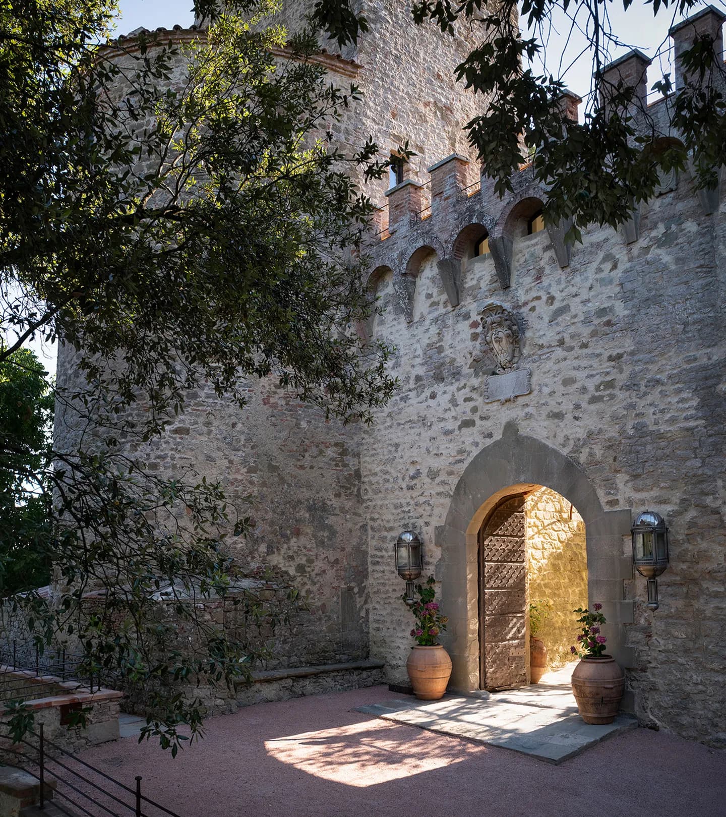 Stone fortress wall with crenellations and arched doorway, crest sculpture above; two large pots flank the entrance as sunlight filters through trees.