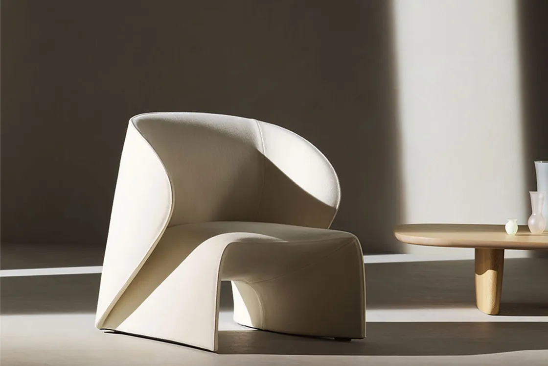 Minimalist cream sculptural chair with sweeping curves beside a round wooden coffee table holding small vases.