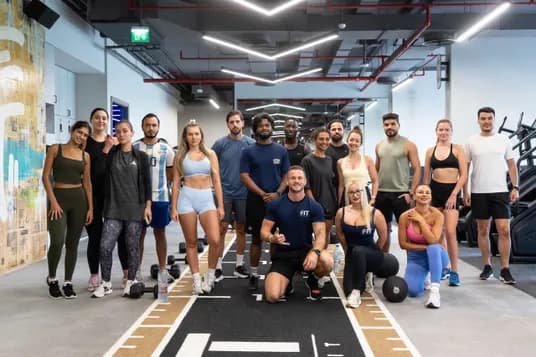 Diverse group of people in a modern gym posing for a photo; trainer kneeling at front center, with weights and gym equipment visible.