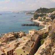 Coastal view of a Mediterranean town with beige buildings, rocky cliffs, and turquoise sea under a partly cloudy sky.