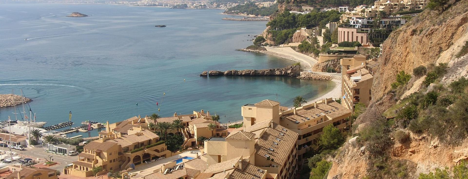 Coastal view of a Mediterranean town with beige buildings, rocky cliffs, and turquoise sea under a partly cloudy sky.