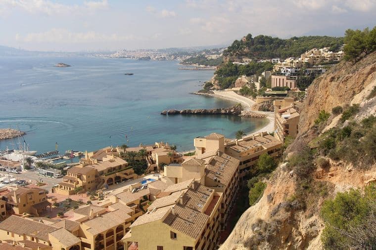 Coastal view of a Mediterranean town with beige buildings, rocky cliffs, and turquoise sea under a partly cloudy sky.