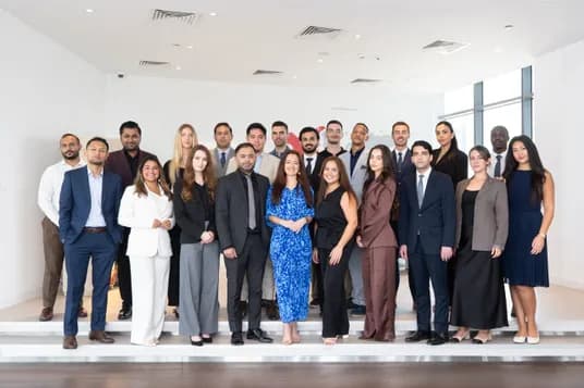 Group of diverse professionals in business attire posing on stairs in a bright, modern office, smiling for a group photo.