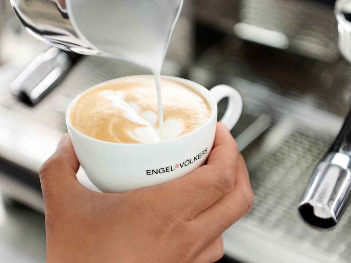 Barista pouring steamed milk into a cappuccino cup, creating latte art. The cup is branded with "ENGEL & VÖLKERS."