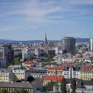 Weitwinkelaufnahme einer europäischen Stadt mit roten Ziegeldächern, einem hohen Kirchturm und modernen Glastürmen vor dem Hintergrund ferner Hügel