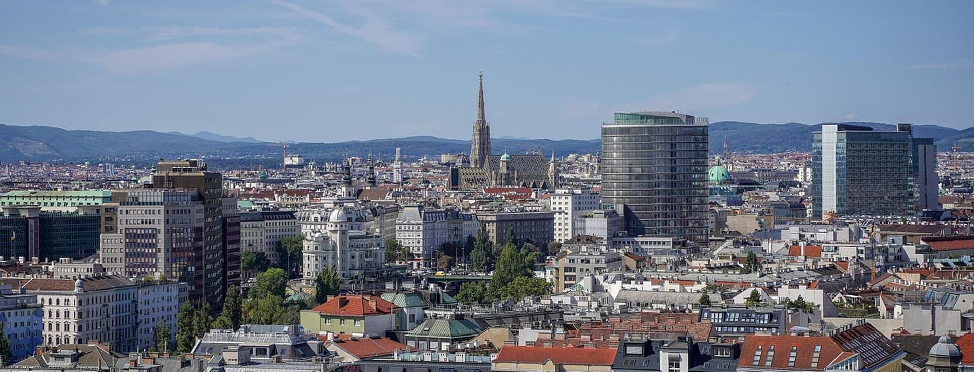 Weitwinkelaufnahme einer europäischen Stadt mit roten Ziegeldächern, einem hohen Kirchturm und modernen Glastürmen vor dem Hintergrund ferner Hügel