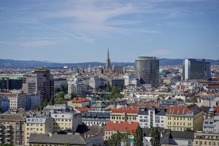 Weitwinkelaufnahme einer europäischen Stadt mit roten Ziegeldächern, einem hohen Kirchturm und modernen Glastürmen vor dem Hintergrund ferner Hügel