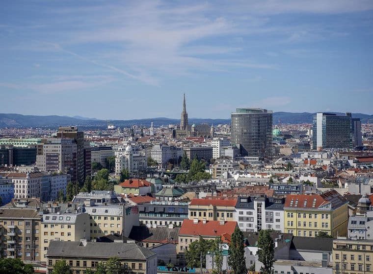 Weitwinkelaufnahme einer europäischen Stadt mit roten Ziegeldächern, einem hohen Kirchturm und modernen Glastürmen vor dem Hintergrund ferner Hügel