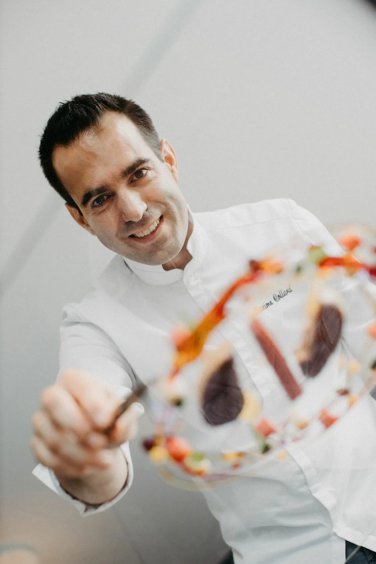 Smiling chef in a white jacket extends toward the camera, presenting a colorful dish in the foreground.