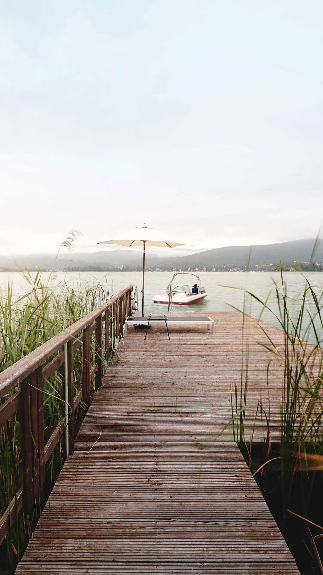 Wooden jetty extends over a lake toward distant hills; a white umbrella shades a lounge chair, while a small boat passes by; reeds frame the sides.