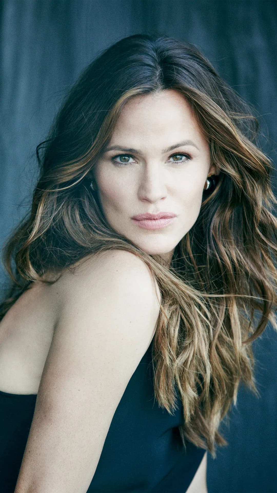Close-up portrait of a woman with long, wavy brunette hair, wearing a dark sleeveless top, gazing at the camera against a dark blue background.
