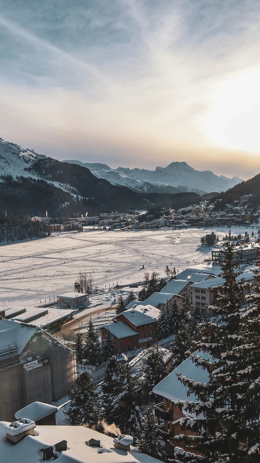 Snowy valley town beneath snow-capped mountains at sunset; ski tracks crisscross the frozen fields framed by pine trees.