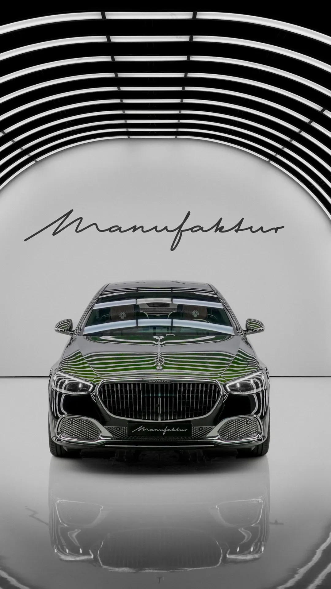 Monochrome front view of a glossy Maybach car with a chrome grille and headlights, reflected on the floor, under curved ceiling arches and 'Manufaktur' on the wall.