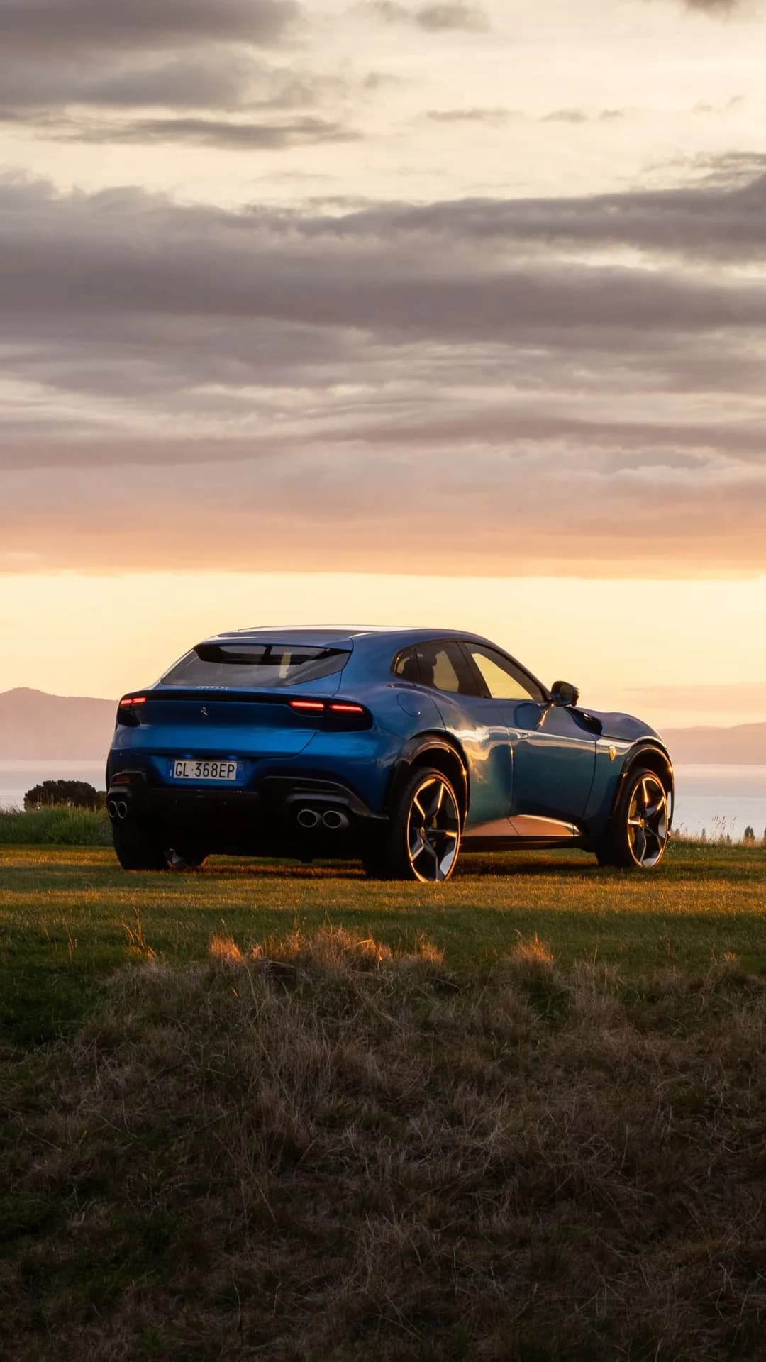 Blue SUV on a grassy hillside at sunset, overlooking a lake with distant mountains and a warm orange sky.