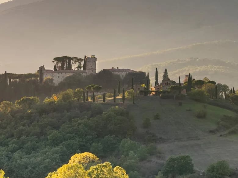 A hilly landscape featuring a castle-like estate with a tall tower, surrounded by cypress trees, set against the backdrop of a warm sunset.