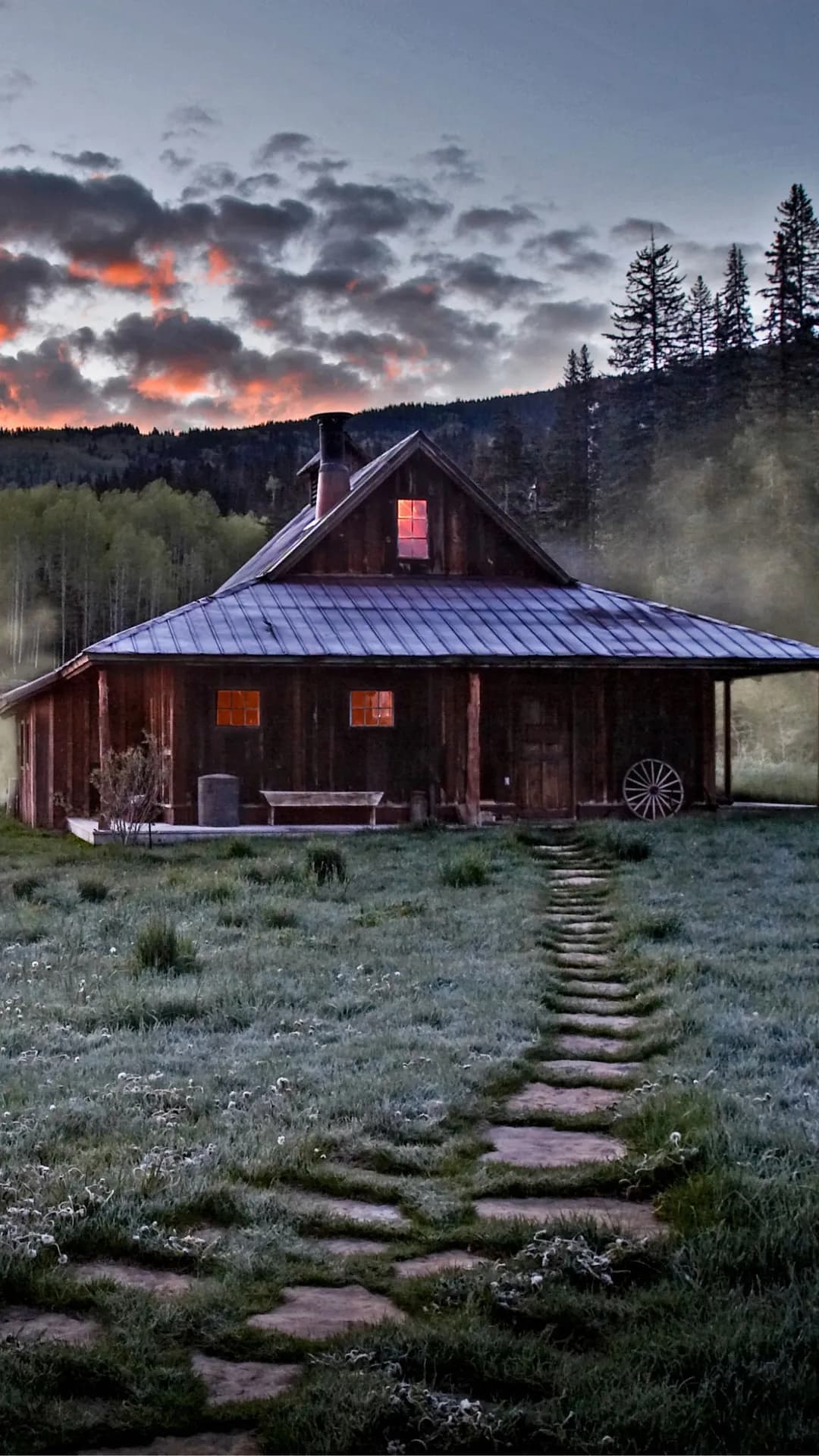Rustic wooden cabin with a metal roof at sunset; glowing orange windows, stone path to the door, wagon wheel beside, forested hills in the background.