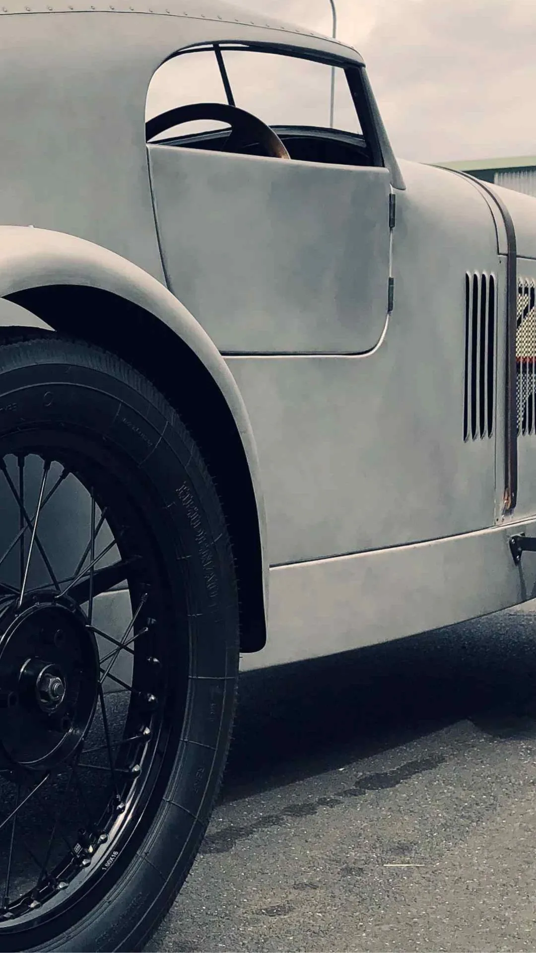 Vintage silver race car with large rounded fenders, wire-spoke wheels, and an open cockpit parked on a paved surface.