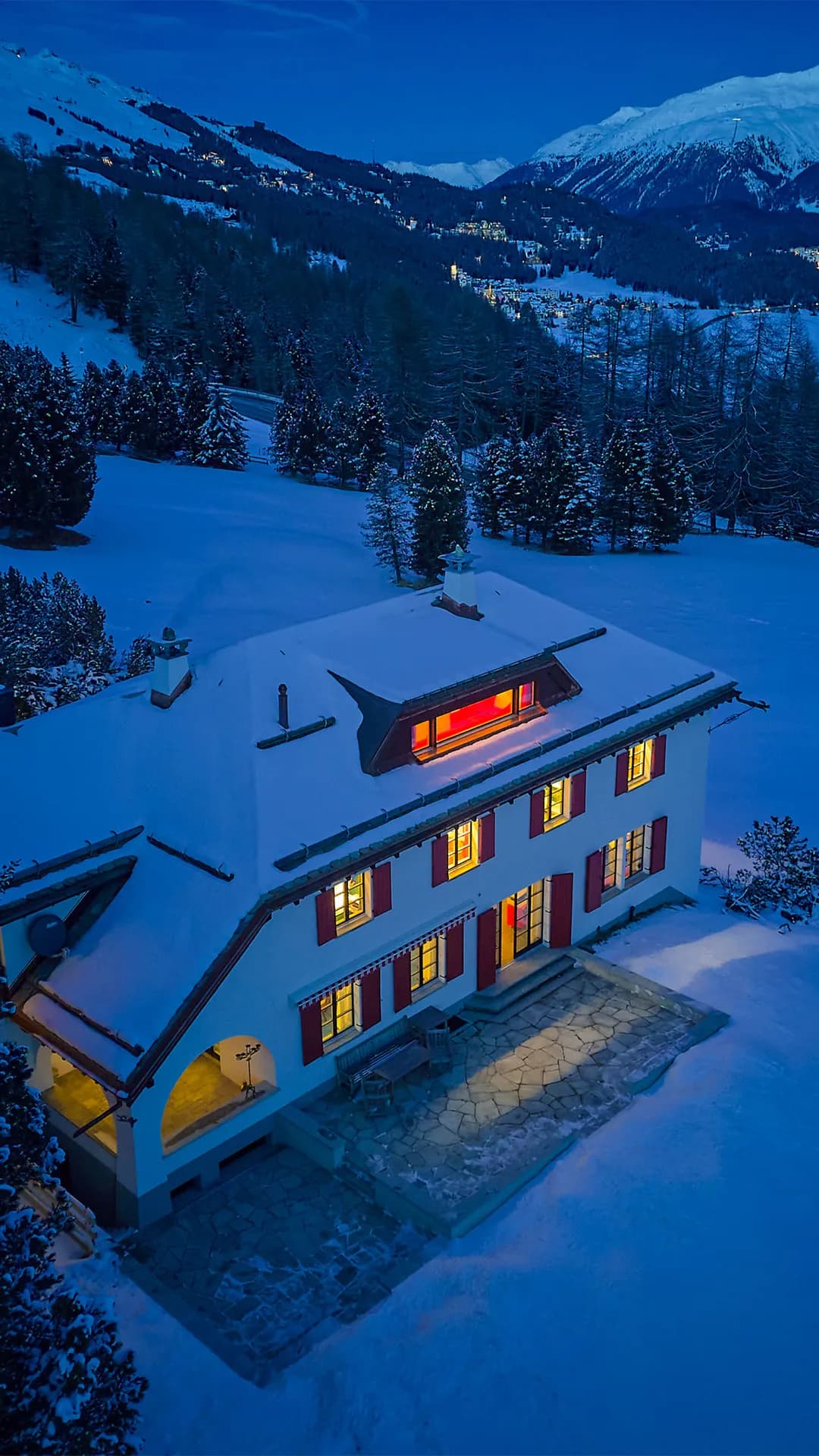 An aerial view of a snow-covered mountain hut surrounded by fir trees, a winding road and snow-covered hills beneath a bright blue sky.