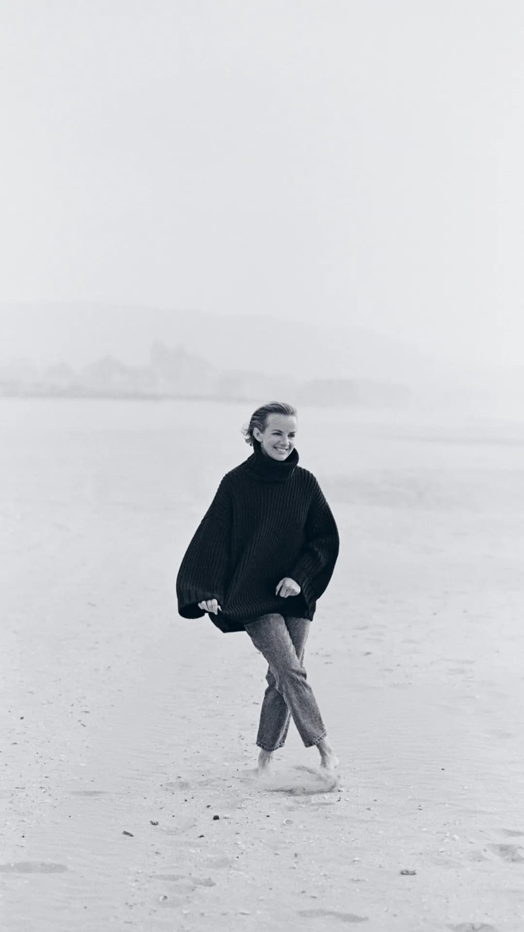 Woman in an oversized black sweater walks barefoot on a misty beach, smiling, with a hazy shoreline visible in the background.