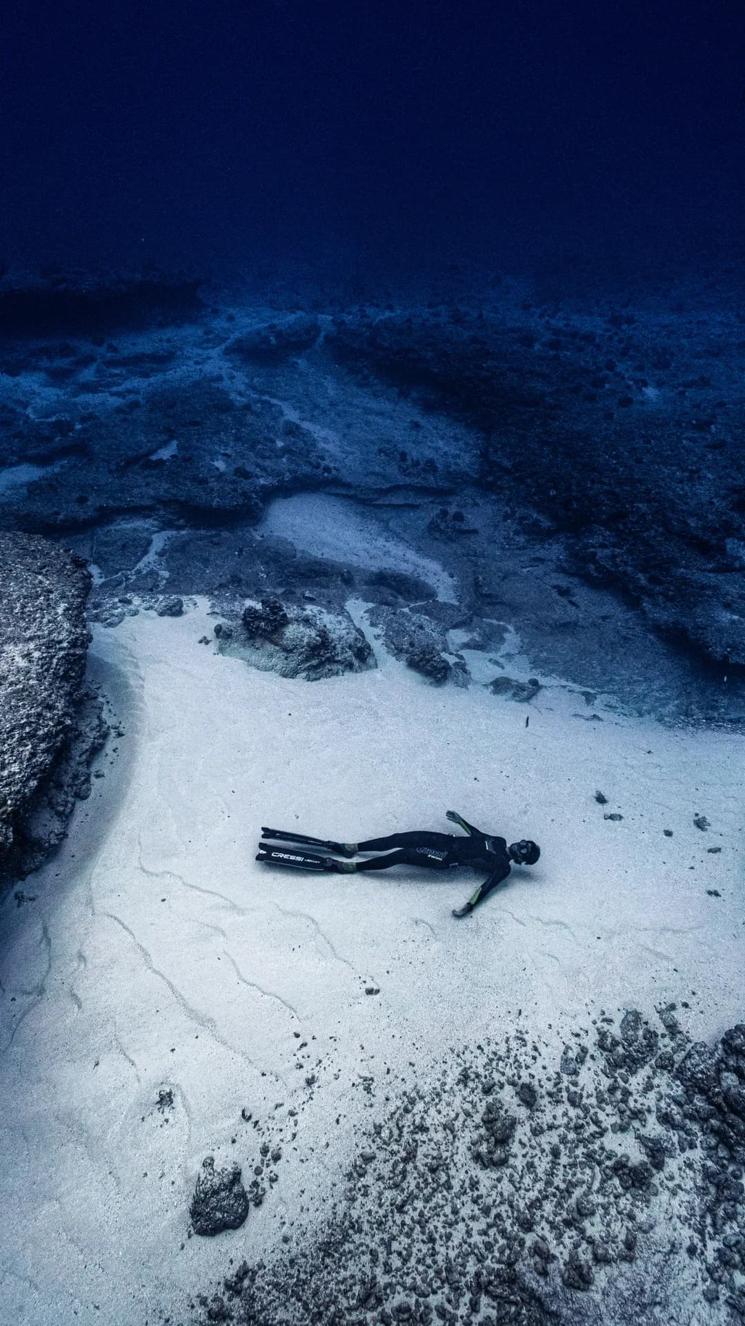 Underwater scene of a scuba diver lying on white sand near rocky outcrops, fins on, as dark blue water looms above.