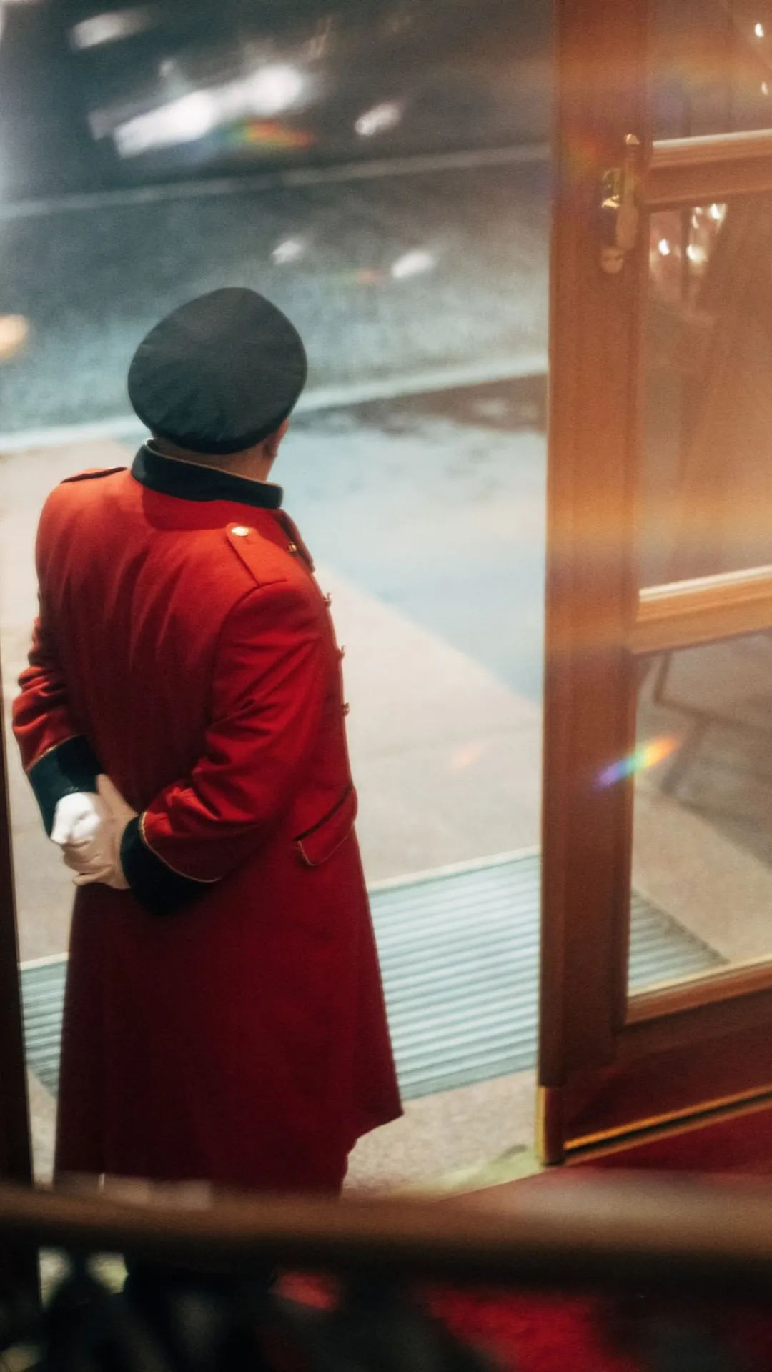 Un fattorino d'albergo con un cappotto rosso e un cappello nero è in piedi davanti a una porta a vetri aperta, con le mani incrociate dietro la schiena, e guarda fuori verso una strada delicatamente illuminata.