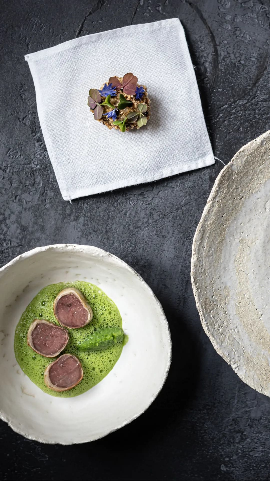 Dark surface with a pale plate holding a chocolate pool topped by a white four-hole button; a white bowl contains green sauce with pink slices; a napkin bears a floral cluster.