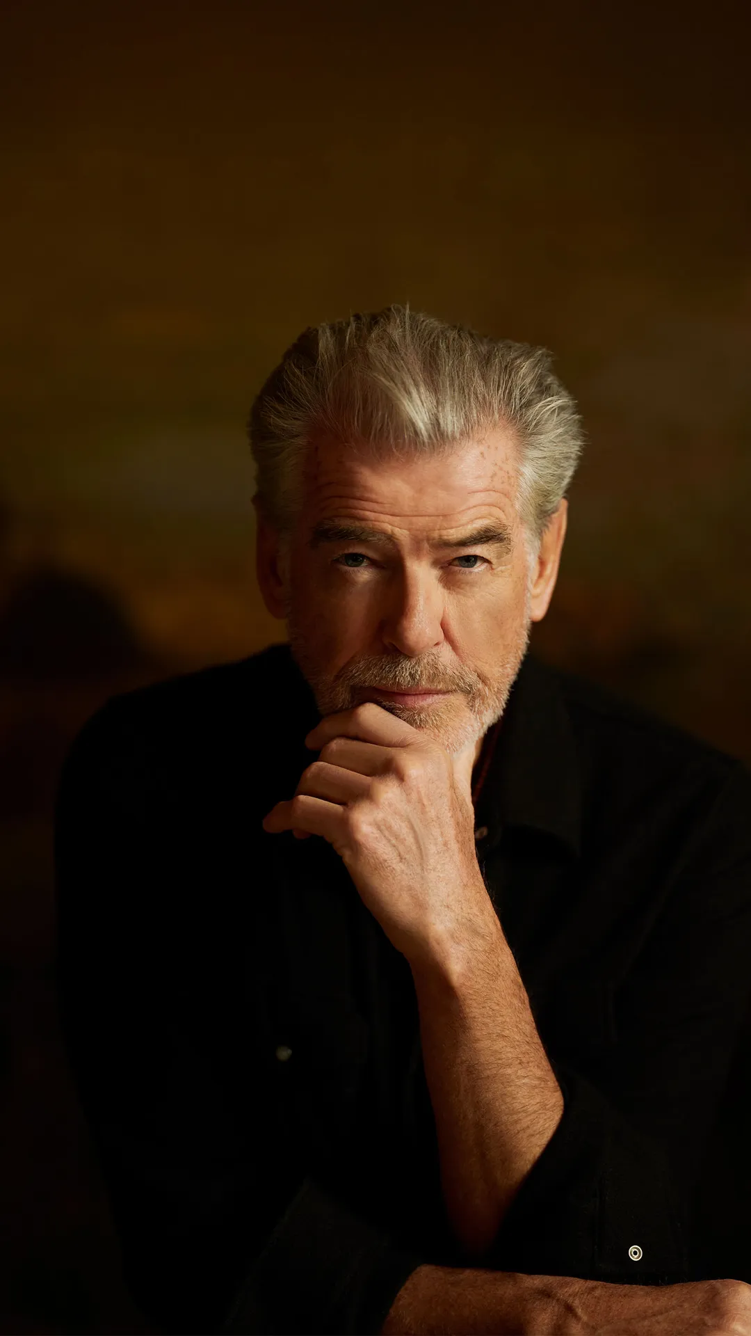 An older man with gray hair and stubble, wearing a black shirt, rests his chin on his hand in a thoughtful pose against a dark, warm background.