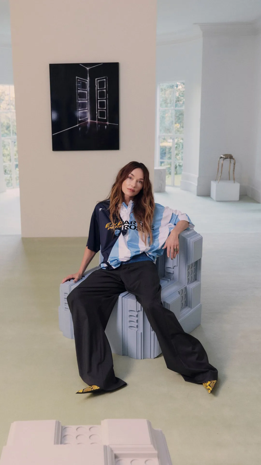 A person is sitting on a blue, sculptural chair in a bright gallery; on the left are light installations, and on the right a wooden chair and display cases.