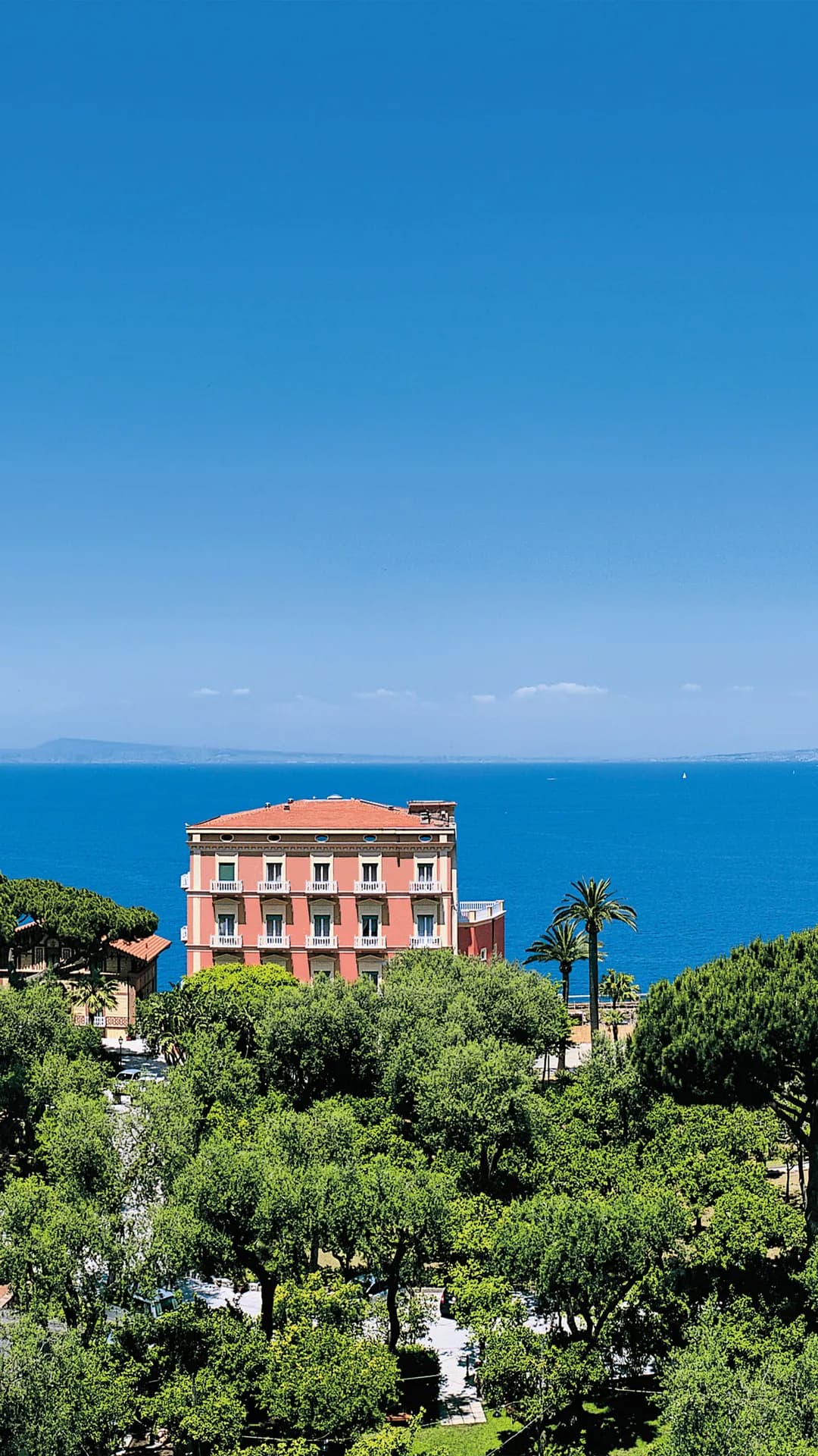 A pink, multi-story seaside building with balconies sits among lush trees, overlooking the blue sea under a clear sky.