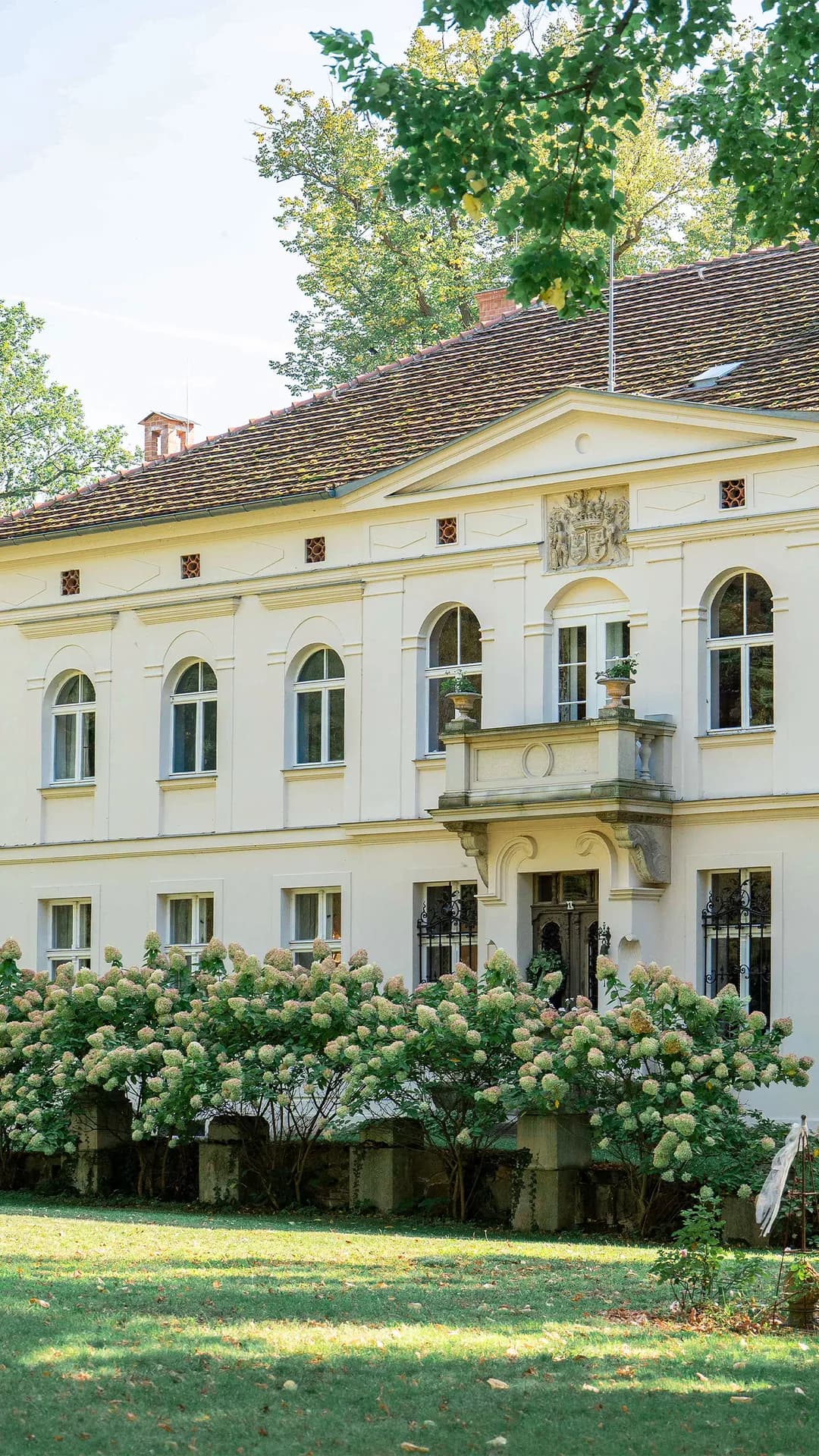 Cream-colored neoclassical building with arched windows, decorative balcony, and crest, shaded by trees, overlooking a garden of hydrangea bushes.