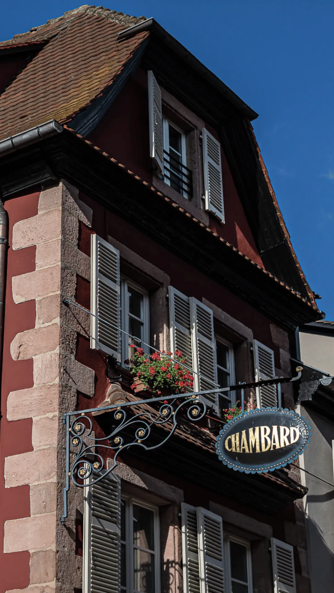 Red brick building with grey shutters, flower boxes on a wrought-iron balcony, and a CHAMBARD sign under a blue sky.