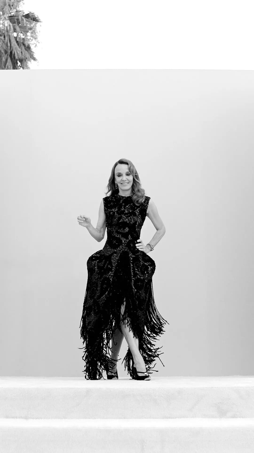 Black-and-white photo of a smiling woman in a long black fringed dress standing on steps outdoors against a pale wall.