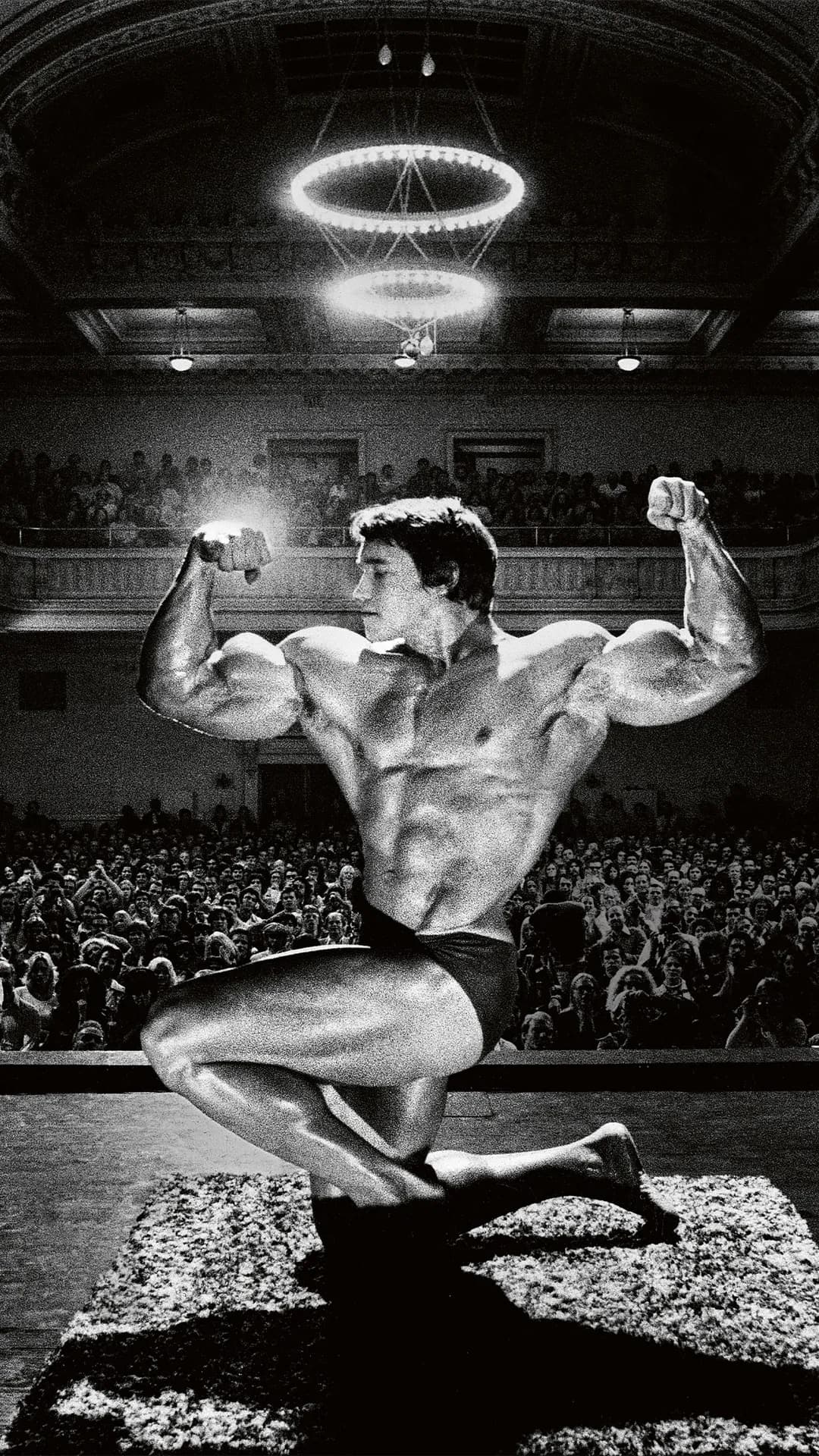 Black-and-white photo of a muscular male bodybuilder on stage, kneeling while flexing his biceps, as a crowd watches from the auditorium.