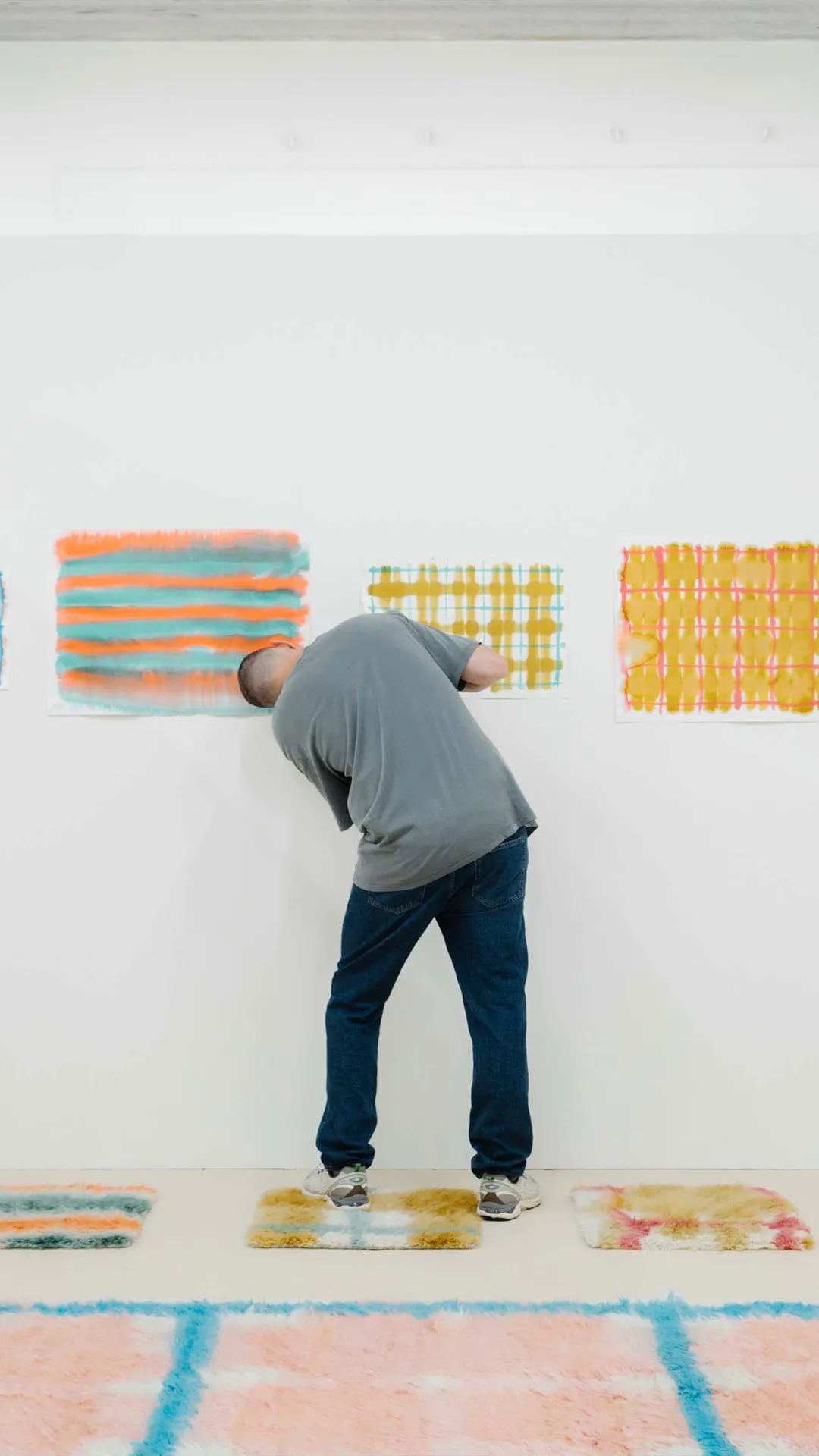 Person in a gray shirt and jeans leans over a white wall, painting bright striped and checkered patterns; colorful square patches on the floor.