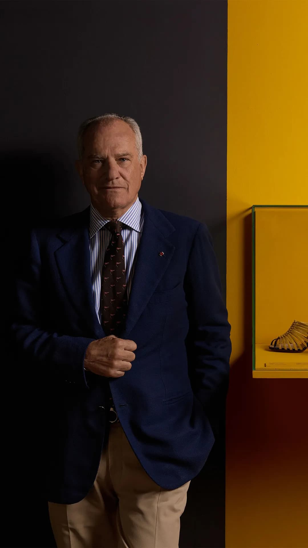Older man in a navy blazer and striped shirt stands beside a yellow display case on a two-tone dark and yellow wall.
